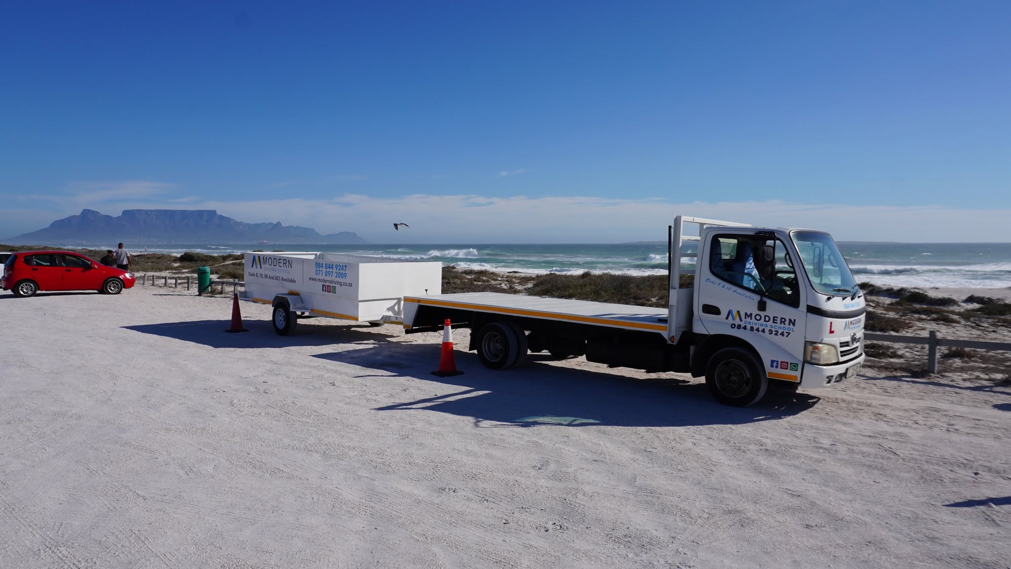 A Modern Driving School code 10 truck and its trailer parked at the coast, with Table Mountain across the bay.